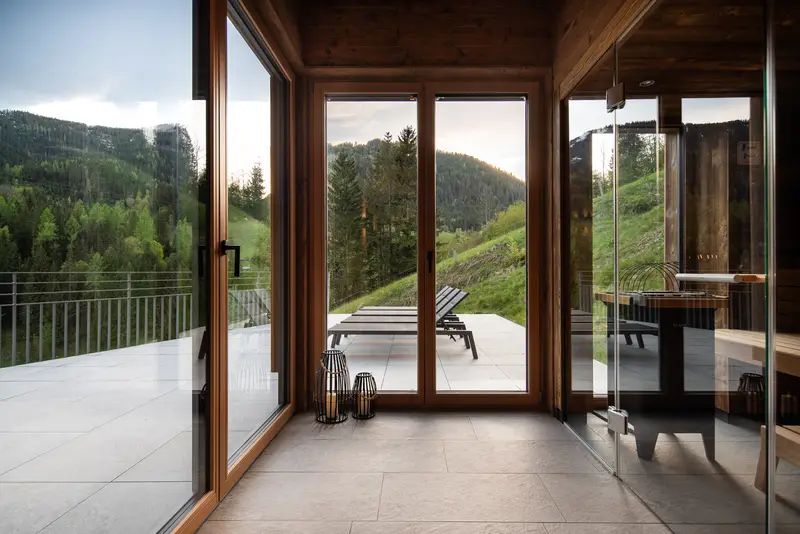Blick durch raumhohe Fenster auf Terrasse, Liegen und grüne Berge